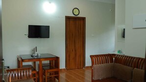 A living room with a wooden dining table, chairs, a sofa, a flat-screen TV mounted on the wall, and a wooden door. - Prakrithi Luxe Retreat (Vythiri)