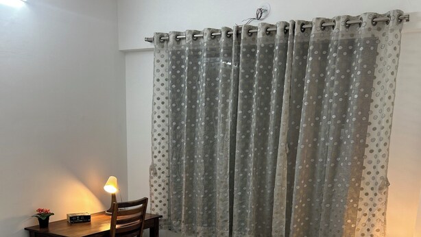 Un dormitorio con cama, escritorio y silla, una mesita de noche con lámpara, y ventana con cortinas.