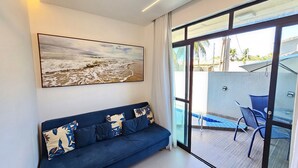 A modern living room with a blue sofa, a large abstract painting, and a glass door leading to an outdoor area with a pool and chairs. - Cupe Summer - Porto de Galinhas (Ipojuca)