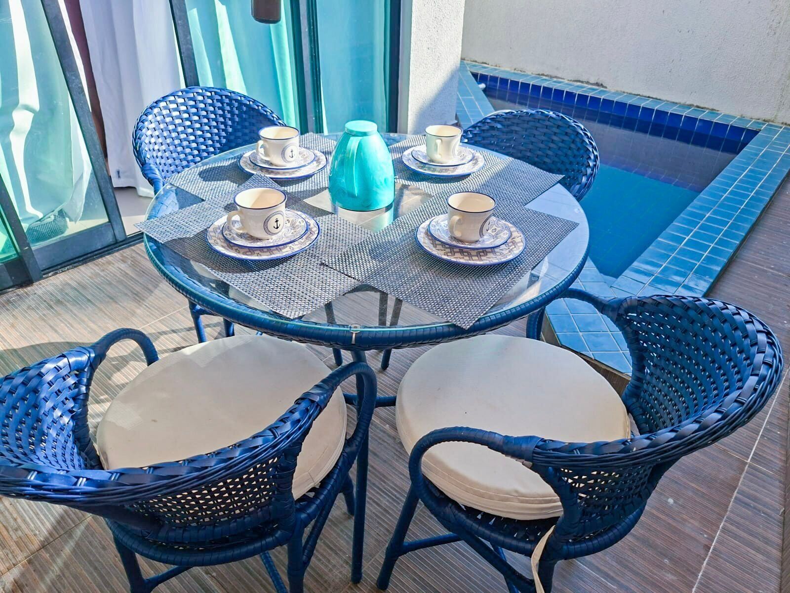 A round outdoor table with four chairs, each with a cushion, and a blue tiled pool adjacent to the table.