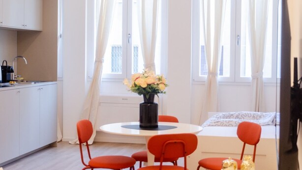 Ein modernes Zimmer mit Esstisch, Stühlen, einer Blumenvase, einem Bett und einem Fernseher.