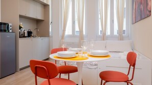 Eine moderne Küche mit weißen Schränken, einem Kühlschrank sowie einem Essbereich mit einem weißen Tisch, orangefarbenen Stühlen und einer Couch.