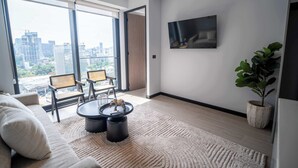 Un salon moderne comprenant un canapé, deux fauteuils, une table basse, un téléviseur et une grande fenêtre donnant sur la ville.