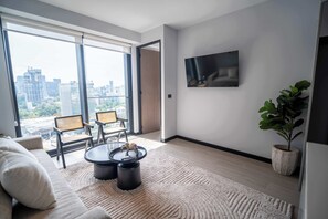 A modern living room with a sofa, two chairs, a coffee table, a TV, and a large window with a city view.