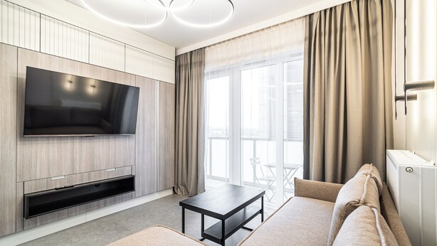 A modern living room with a flat-screen TV, a sofa, a coffee table, and a large window with curtains. - RentPlanet - Studio Zabrska (Katowice)