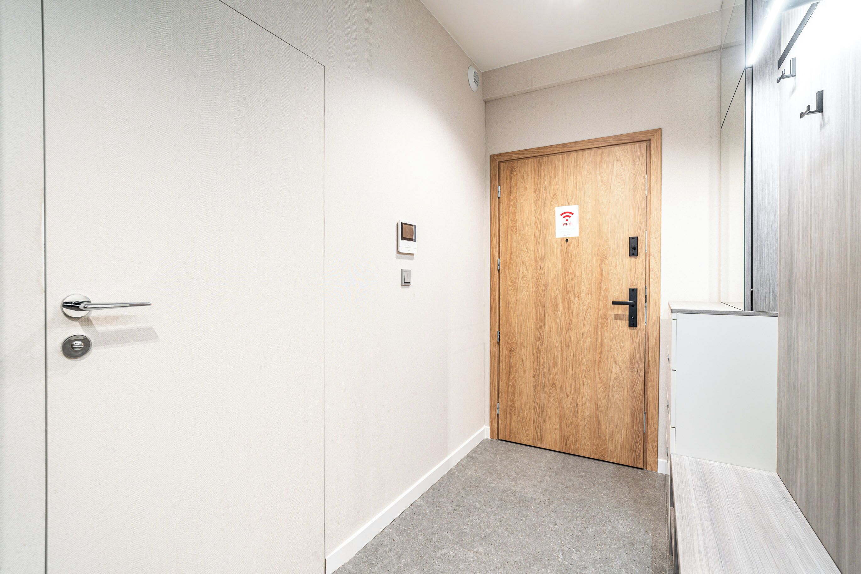 A hallway with a wooden door, a white wall, and a light-colored floor.