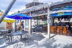 An outdoor bar area with tiki-style umbrellas, metal chairs, and a bar counter. - Purple Parrot Condo Near Beach 2 Bedroom (pensacola)