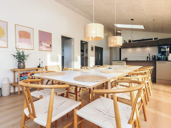 A spacious dining area with wooden chairs and tables, a kitchen in the background, and pendant lighting.