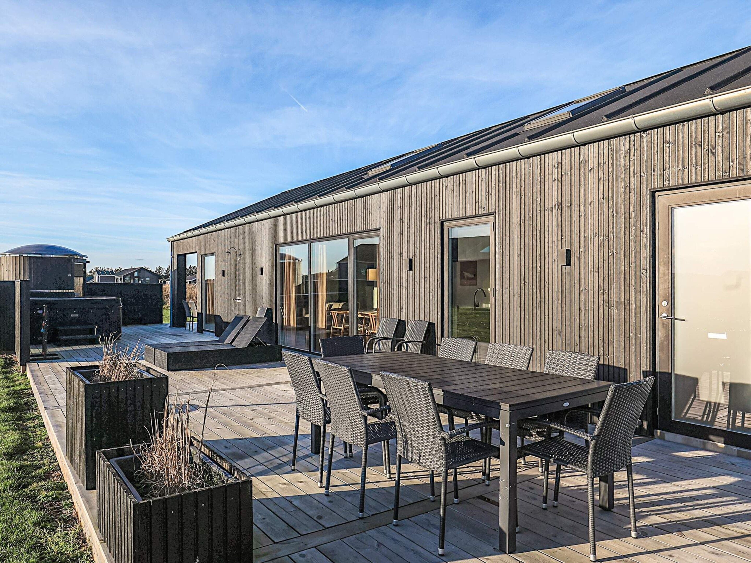 A modern outdoor patio with a wooden deck, a dining table with chairs, and a large window overlooking a grassy area.