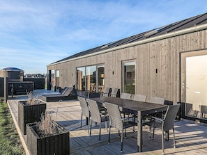 A modern outdoor patio with a wooden deck, a dining table with chairs, and a large window overlooking a grassy area.