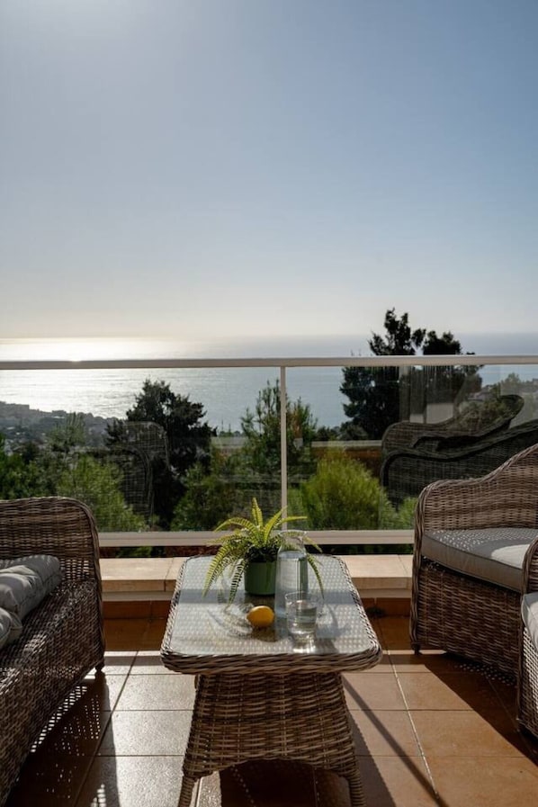 A balcony with wicker furniture, a glass table, and a view of the sea.
