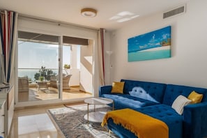 A living room with a blue sofa, a glass door leading to a balcony, a painting of a beach scene, and a patterned rug.
