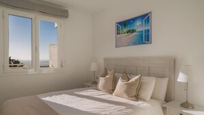 A bedroom with a bed, two bedside tables, a nightstand, a window, and a painting of a beach scene. - Stylish 2BR Apt | Balcony & Sea Views (Benalmádena)