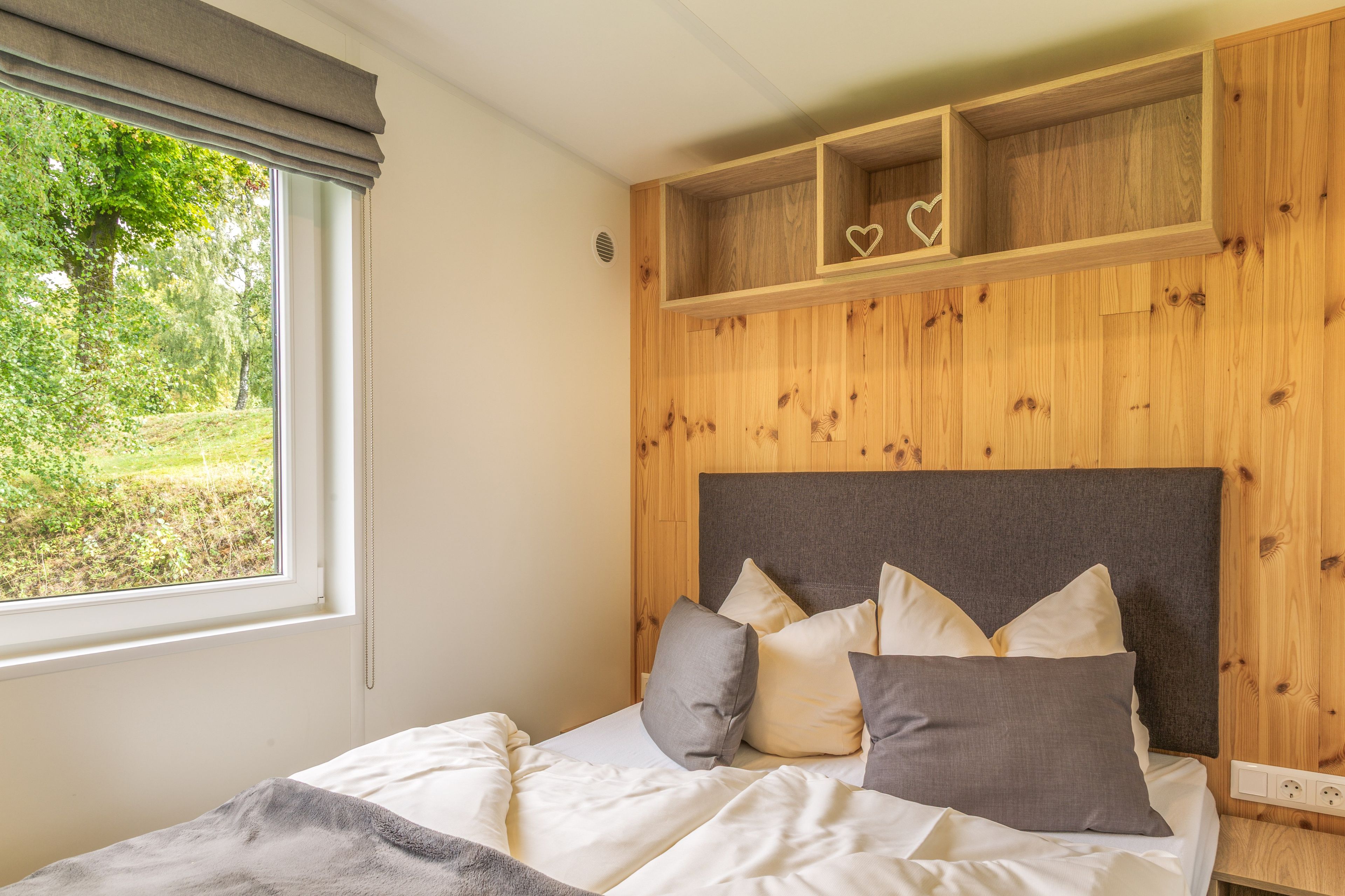 A bedroom with a bed, pillows, a window with curtains, and a wooden headboard with a shelf above.