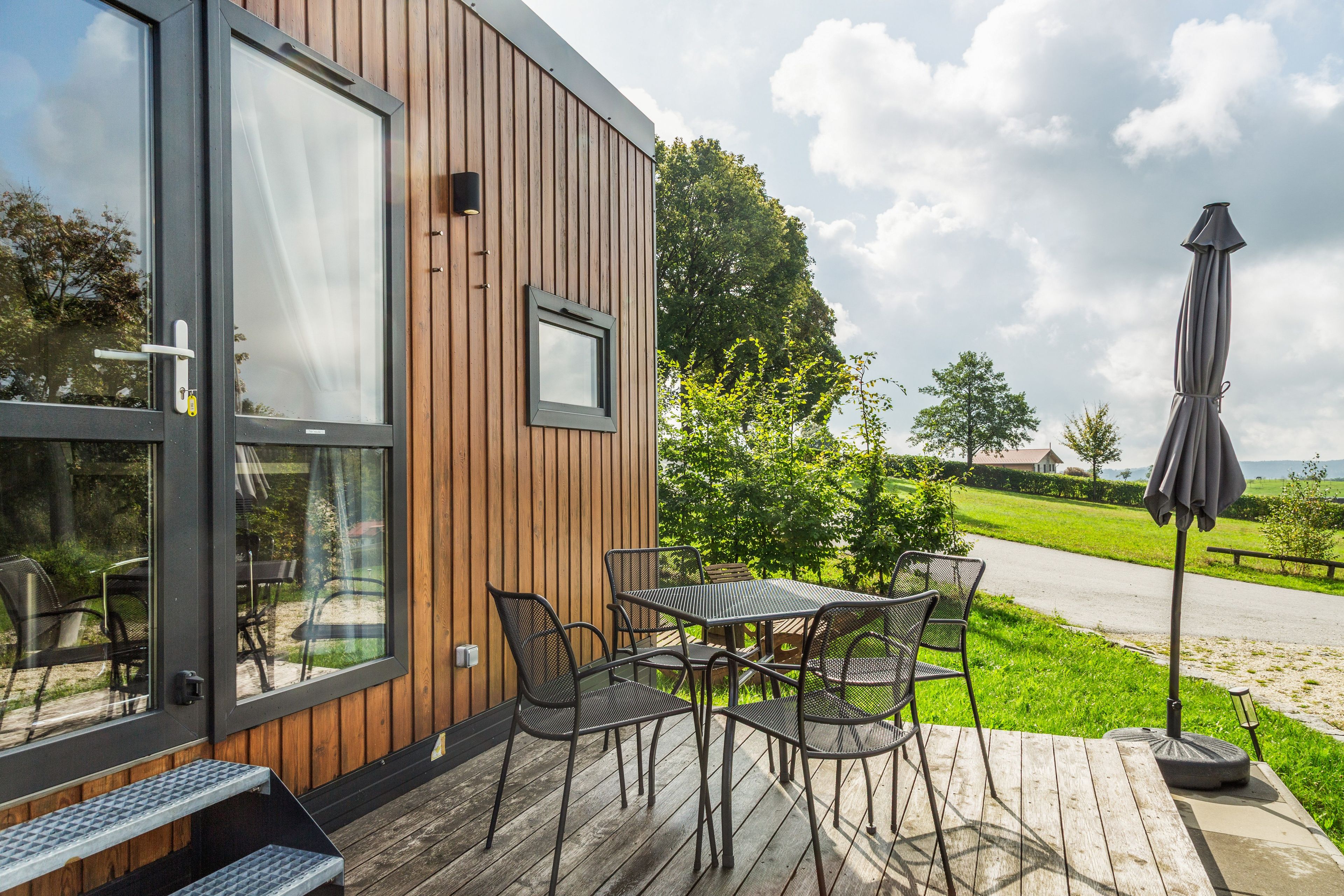 Eine Holzfassade mit Glastür und -fenster, Sitzgelegenheiten im Freien mit Metallstühlen und einem Tisch, sowie eine Rasenfläche mit einem Weg und Bäumen.