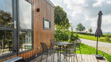 Eine Holzfassade mit Glastür und -fenster, Sitzgelegenheiten im Freien mit Metallstühlen und einem Tisch, sowie eine Rasenfläche mit einem Weg und Bäumen.