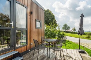 A wooden exterior wall with a glass door and window, outdoor seating with metal chairs and a table, and a grassy area with a path and trees. - Tiny Home 'Laura' with Mountain Views, Wi-Fi, and Air Conditioning (Untergriesbach)