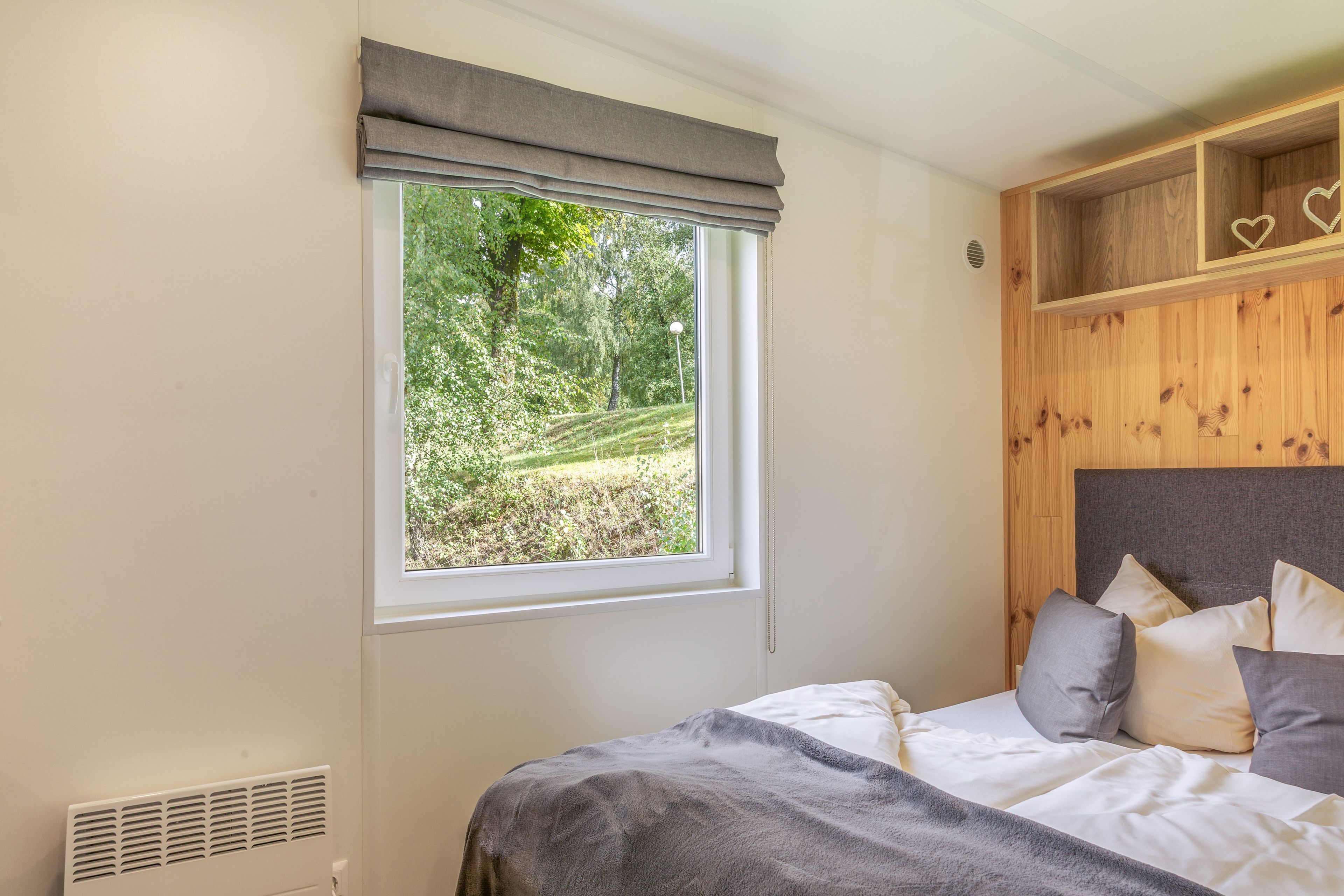 A bedroom with a bed, a window with a view of greenery, a wooden headboard, and a shelf with heart-shaped decorations.