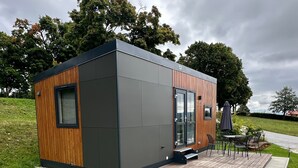 A modern, compact building with a wooden exterior, a glass door entrance, and a small outdoor seating area. - Tiny Home 'Laura' with Mountain Views, Wi-Fi, and Air Conditioning (Untergriesbach)