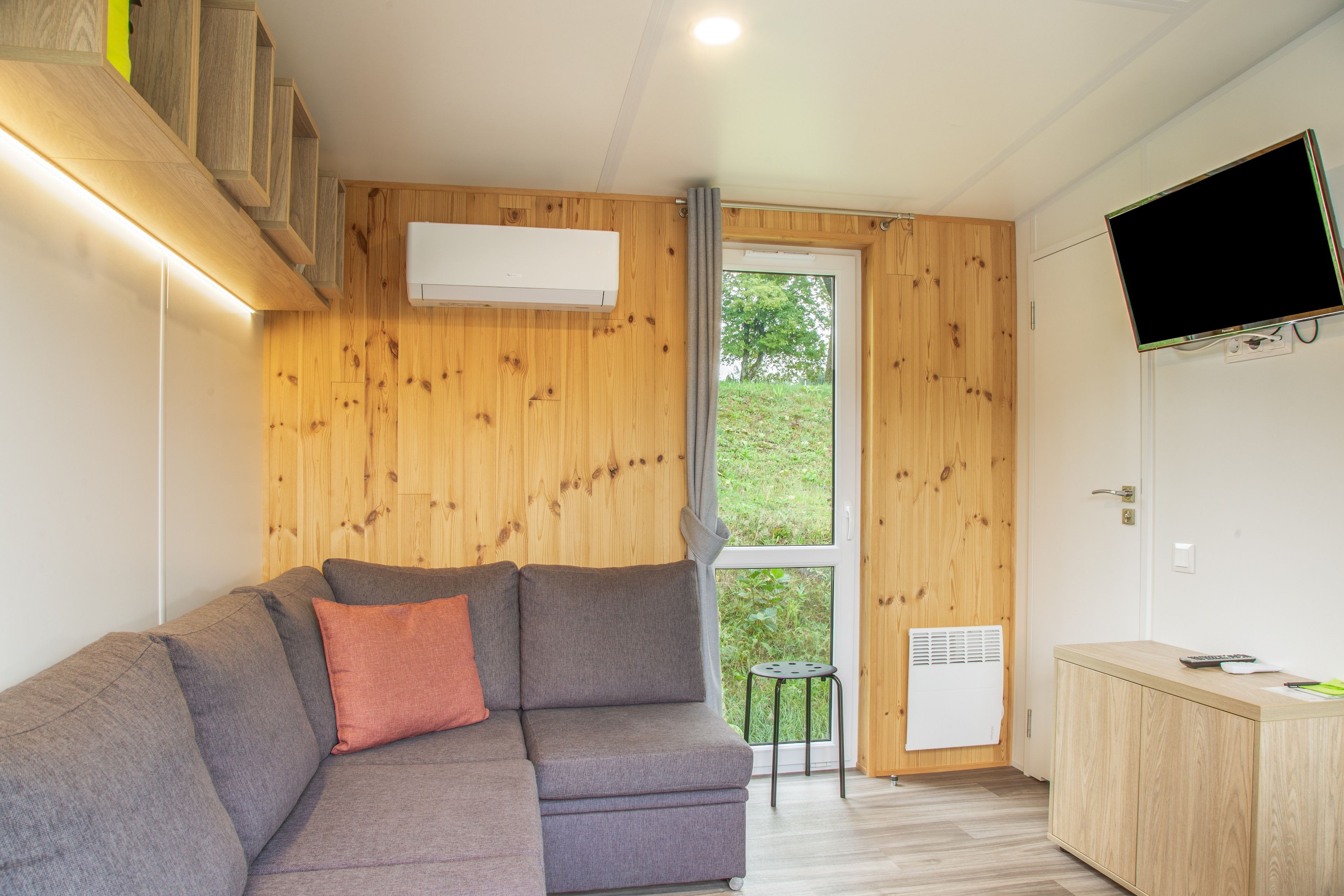 A modern interior with a grey sofa, wooden walls, a TV, and a window with a view of greenery.