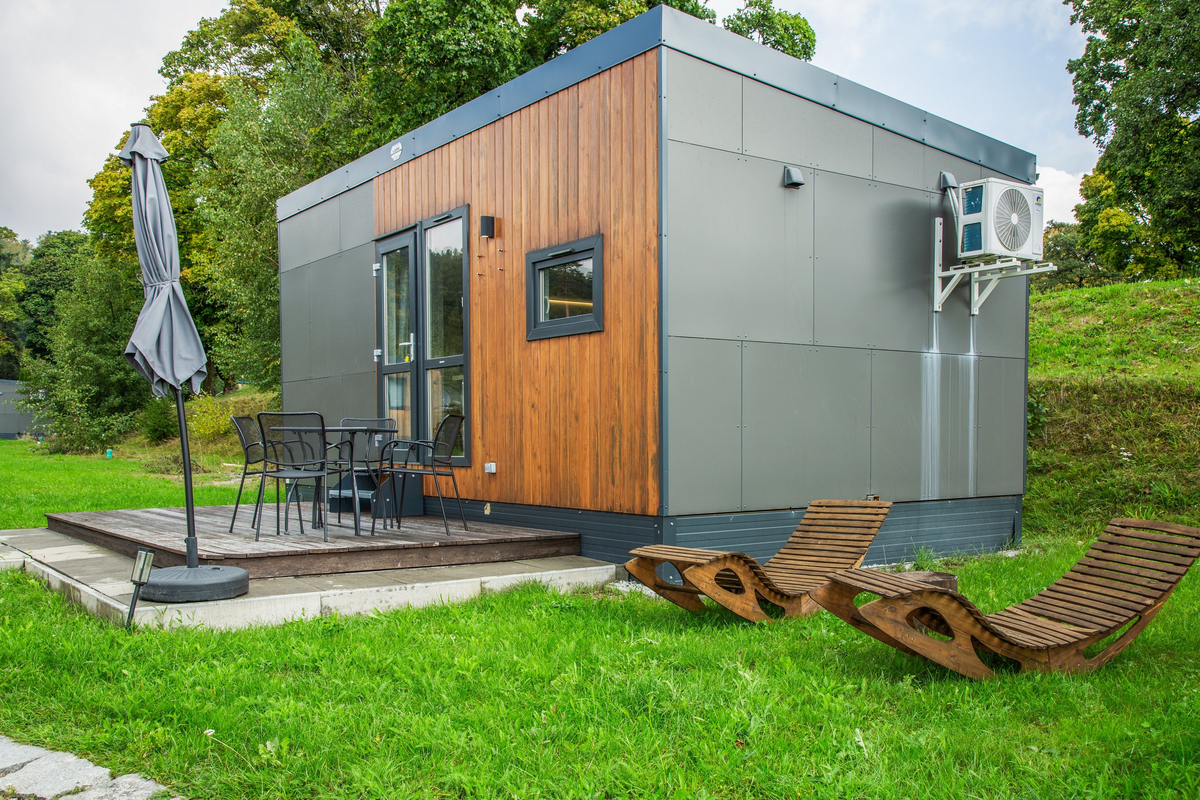 Ein modernes, kompaktes Gebäude mit Holzfassade, einer Terrasse und Gartenmöbeln sowie einer an der Seite angebrachten Klimaanlage.
