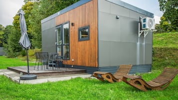 Ein modernes, kompaktes Gebäude mit Holzfassade, einer Terrasse und Gartenmöbeln sowie einer an der Seite angebrachten Klimaanlage.