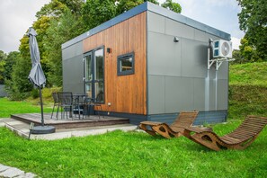 Un edificio moderno y compacto con fachada de madera, un patio con mobiliario exterior y una unidad de aire acondicionado en el lateral.