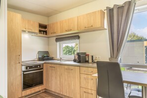 A compact kitchen with wooden cabinets, a built-in oven, a coffee maker, and a window with a view of greenery. - Tiny Home 'Laura' with Mountain Views, Wi-Fi, and Air Conditioning (Untergriesbach)