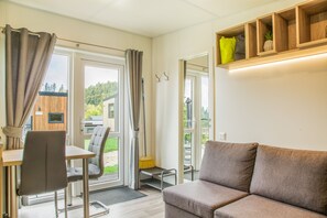 A modern interior with a sofa, a dining table, and a chair. There is a window with curtains, a door, and a small shelf with items. - Tiny Home 'Laura' with Mountain Views, Wi-Fi, and Air Conditioning (Untergriesbach)