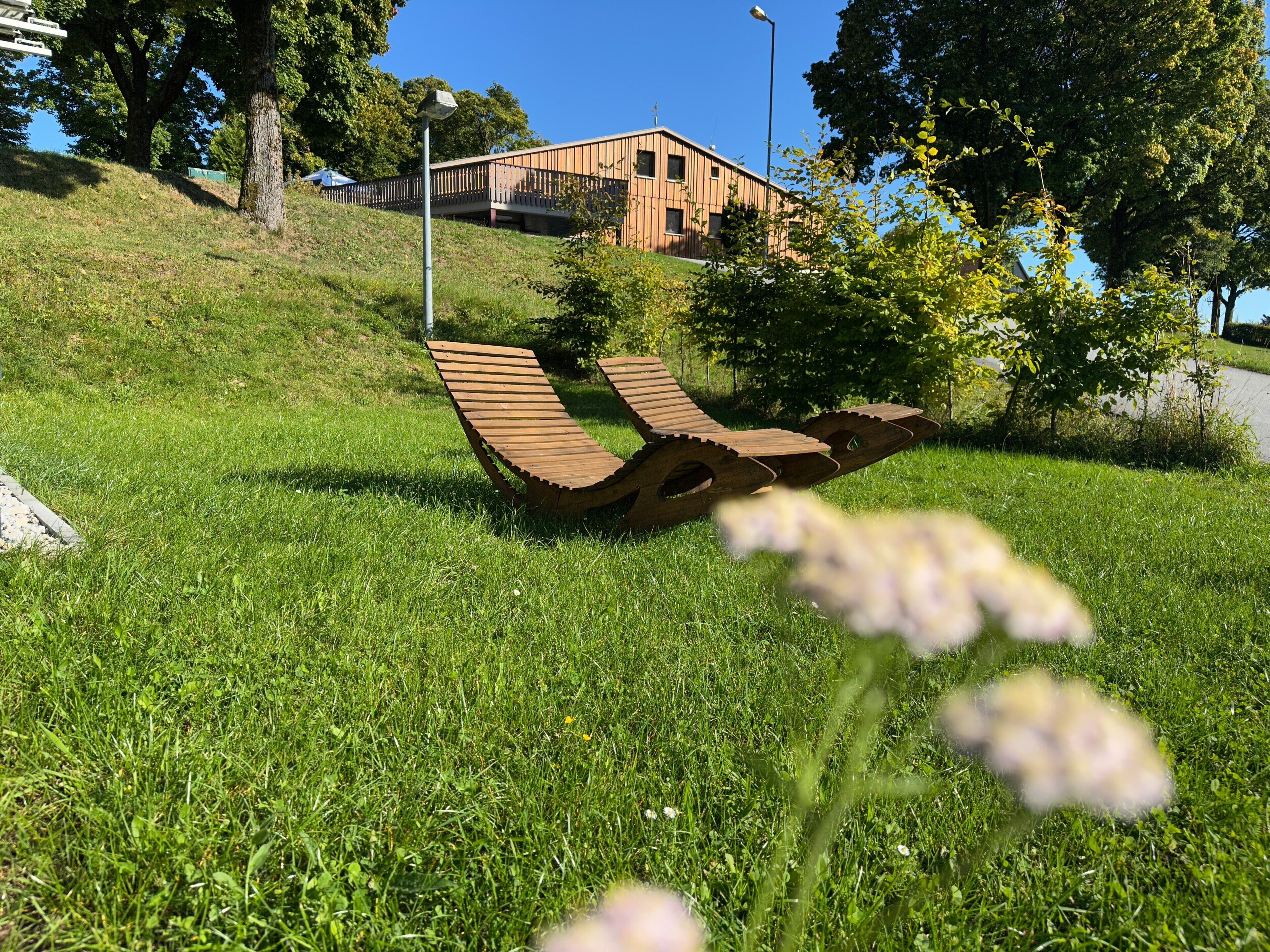 Ein grünes Feld mit einer ungewöhnlichen Holzbank-Konstruktion, einem Gebäude im Hintergrund und einem strahlend blauen Himmel.