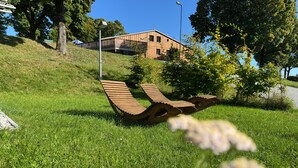 Un espace verdoyant agrémenté d’un banc en bois à la structure originale, un bâtiment en arrière-plan et un ciel bleu dégagé.
