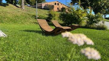 Ein grünes Feld mit einer ungewöhnlichen Holzbank-Konstruktion, einem Gebäude im Hintergrund und einem strahlend blauen Himmel.