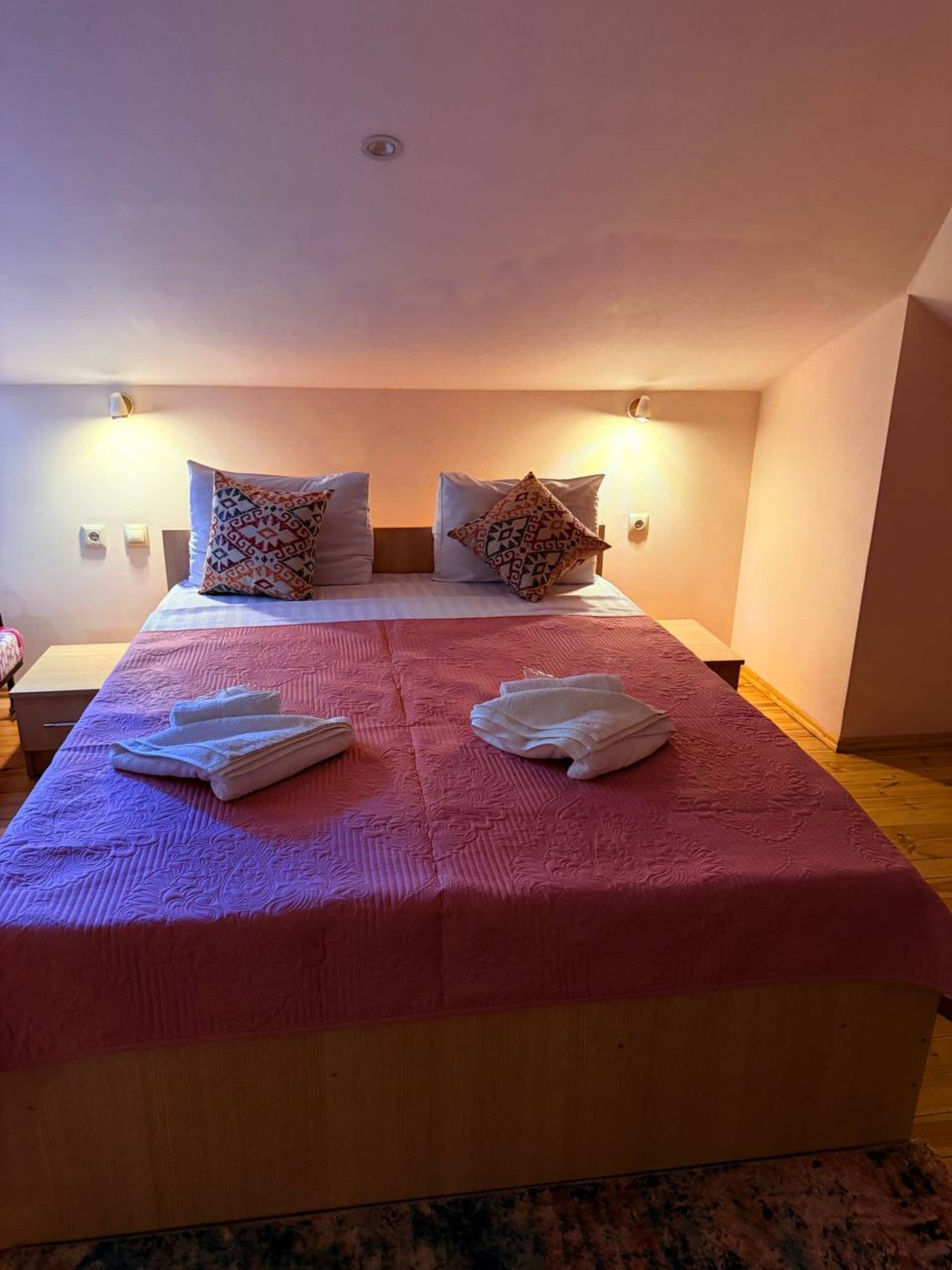 A double bed with patterned pillows, a pink bedspread, and two folded towels on the bed.