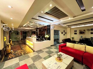Un hall de hotel con mostrador de recepción, un sofá rojo, una mesa de centro con periódicos y un suelo a cuadros.