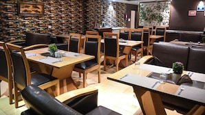 A restaurant with wooden tables and black leather chairs, a stone wall, and a small plant on the tables. - V Hotel TSSG (New Delhi)