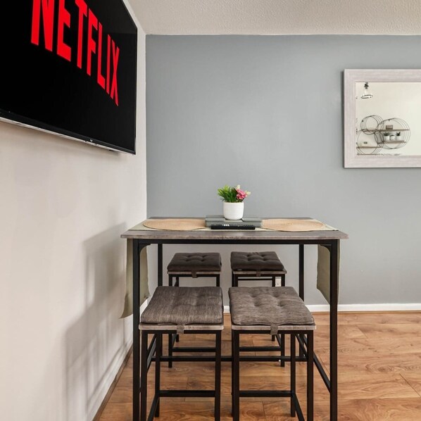 Ein modernes Wohnzimmer mit einem an der Wand befestigten Flachbildfernseher, der 'NETFLIX' anzeigt. Ein kleiner Tisch mit einem Topf und einer Zimmerpflanze steht dort, daneben ein gerahmtes Bild an der Wand. Zwei gepolsterte Hocker sind vor dem Tisch platziert.