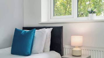 A bedroom with a bed, pillows, a bedside table with a lamp, and a window with a view of trees.