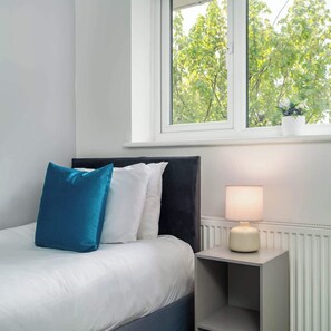 A bedroom with a bed, pillows, a bedside table with a lamp, and a window with a view of trees.