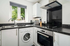 A compact kitchen with a washing machine, oven, and a window with a view of greenery.