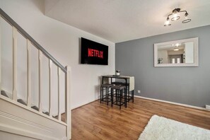 A modern living room with a wooden floor, a white staircase, a small table with two chairs, a flat-screen TV mounted on the wall, and a mirror above the table.