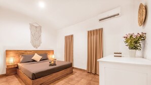 Habitación de hotel con una cama de madera, dos lámparas de noche, un aire acondicionado de pared y una pequeña mesa blanca con un jarrón de flores.