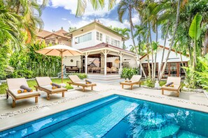 Un espace aménagé au bord de la piscine, avec des chaises longues et des parasols, une piscine et une maison dotée d’un toit en tuiles et de grandes fenêtres.
