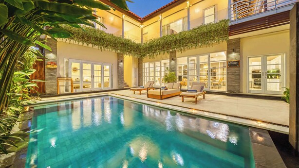A swimming pool with lounge chairs and a patio area, surrounded by a building with large windows and a balcony.