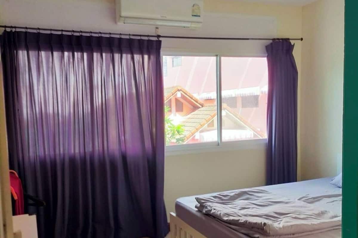 A bedroom with a bed, a window with curtains, an air conditioning unit, and a view of buildings outside.