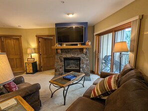 A cozy living room with a stone fireplace, a flat-screen TV on a wooden shelf, leather sofas, and a dining area with a table and chairs.