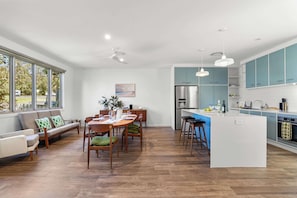 Uma cozinha moderna com uma ilha central, uma sala de jantar com mesa e cadeiras de madeira, e uma sala de estar com sofá e uma janela grande.