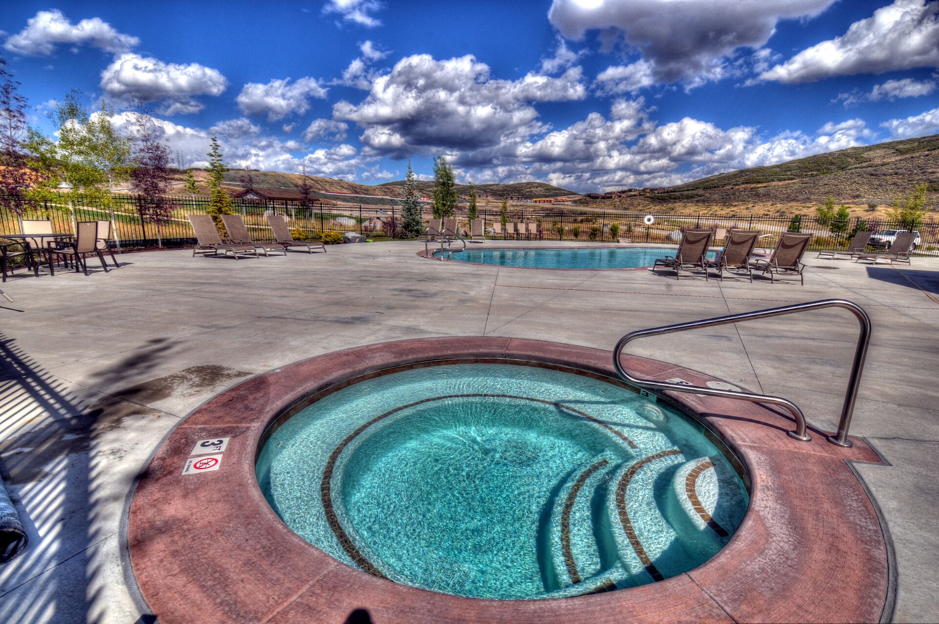 A hot tub surrounded by lounge chairs and a pool, with a mountainous backdrop.