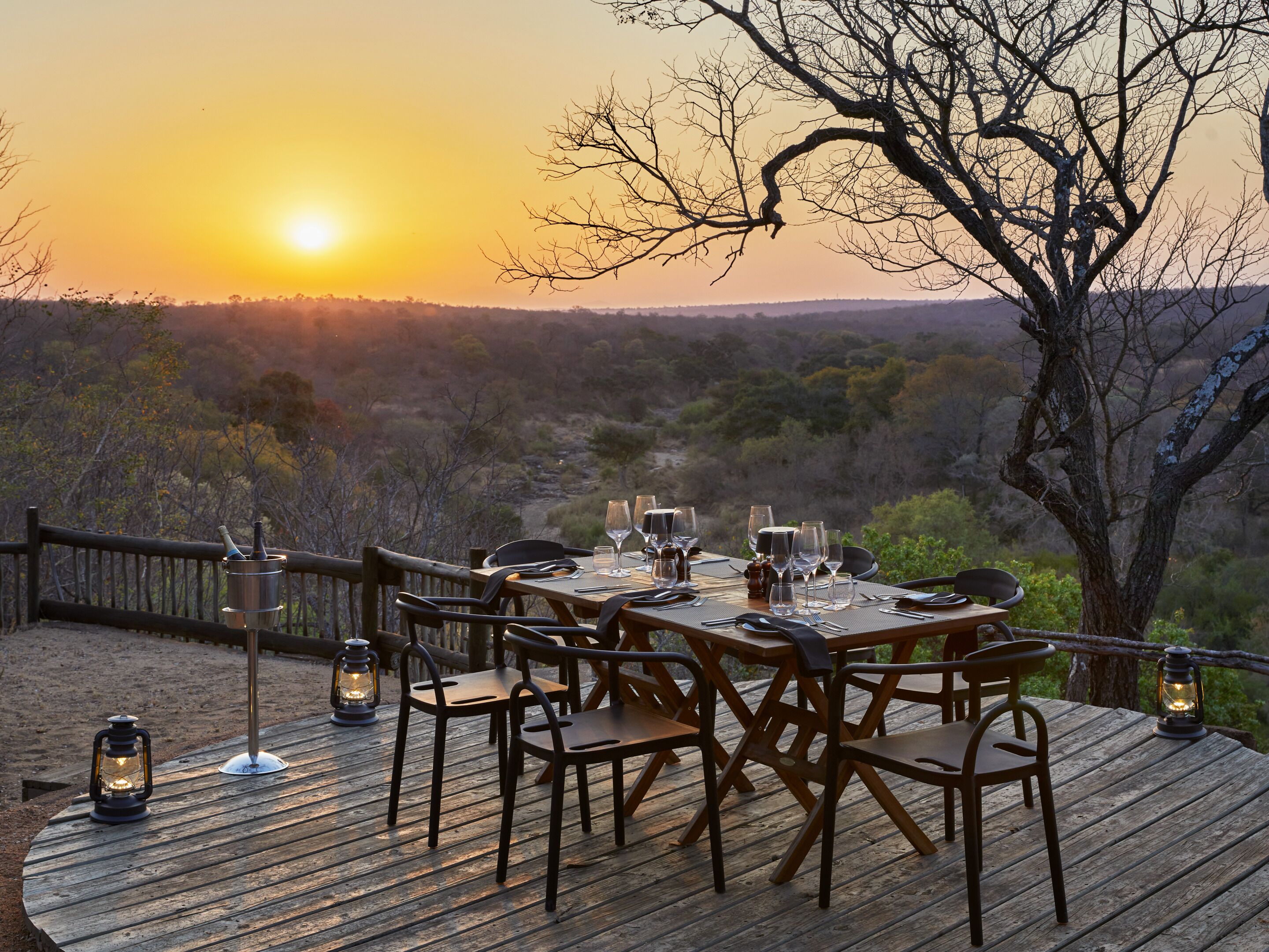 An outdoor dining area with a table set for six, surrounded by chairs, overlooking a scenic landscape during sunset.