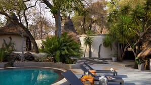 A poolside area with lounge chairs, a small table with drinks, and a thatched-roof structure. - Toro River by Newmark (Makalali Game Reserve)