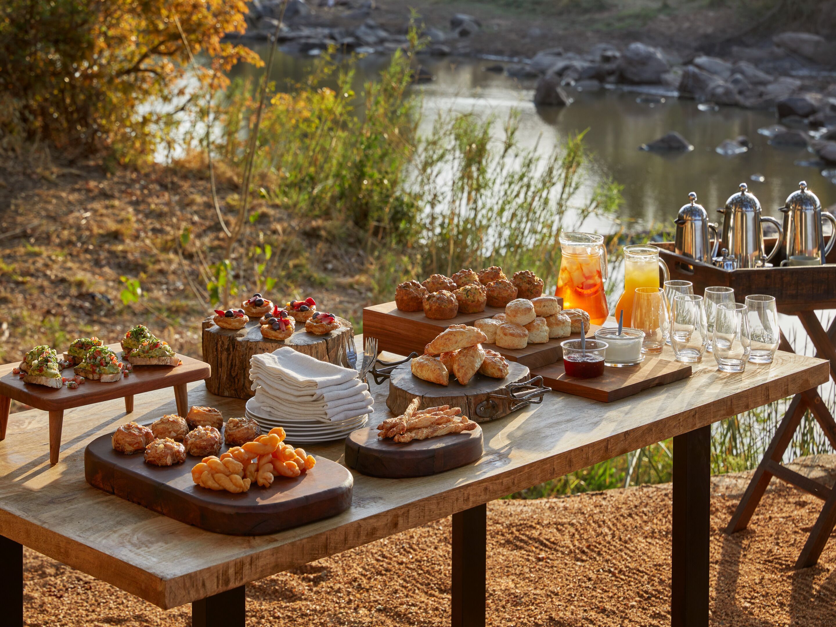 Un pique-nique est installé sur une table en bois près d’une rivière, avec divers aliments et une boisson.